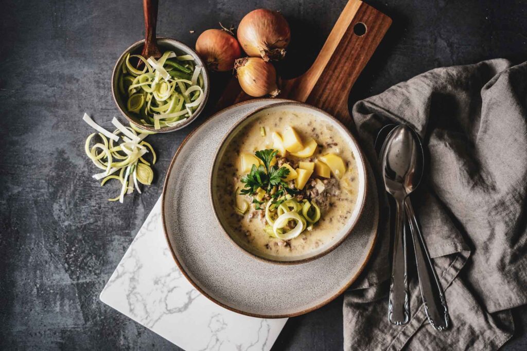 Cremige Käse-Lauch-Suppe mit Hackfleisch und Kartoffeln in einer Steingutschüssel auf einem Holzbrett. Daneben liegen eine graue Stoffserviette, braune Zwiebeln und geschnittene Lauchringe