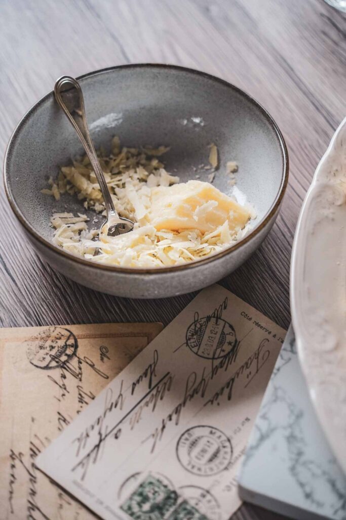 Steingutschüssel mit gehobeltem Parmesan auf einem Holztisch neben Vintage-Postkarten. Detailaufnahme einer typischen Zutat der italienischen Küche für Pasta- und Genussrezepte