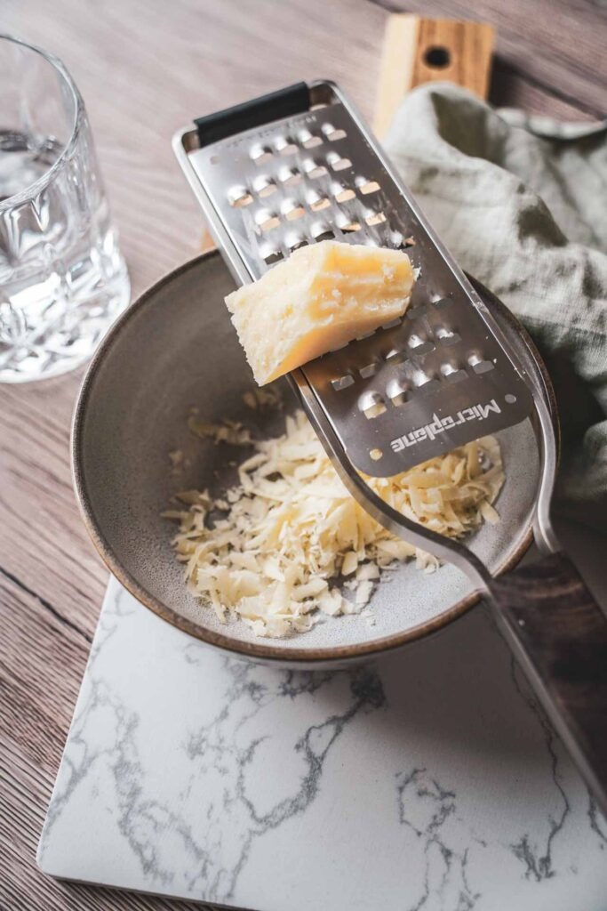 Frisch gehobelter Parmesan in einer Steingutschüssel mit einer Microplane Parmesanreibe auf einem Holzbrett mit hellgrüner Serviette – typische Zutaten für authentische italienische Pasta-Rezepte aus Apulien
