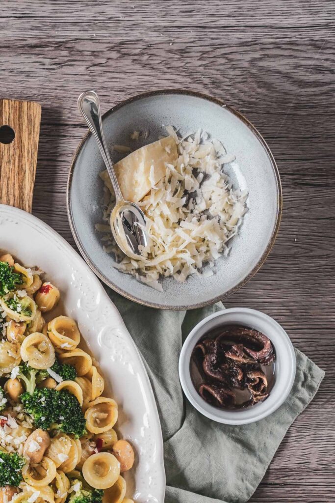 Traditionelle Orecchiette mit Brokkoli und weißen Bohnen aus Apulien auf einer weißen Servierplatte. Das italienische Pastagericht wird von gehobeltem Parmesan, Sardellen in Öl, einer hellgrünen Serviette und Vintage-Postkarten begleitet und erinnert an die mediterrane Küche Süditaliens