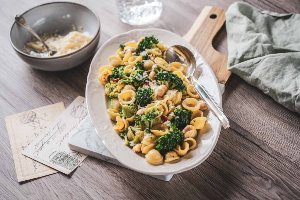 Traditionelles Orecchiette-Rezept aus Apulien mit Brokkoli und weißen Bohnen. Die italienische Pasta wird auf einer weißen Servierplatte angerichtet und mit Parmesan, Vintage-Postkarten und mediterranen Details stilvoll in Szene gesetzt