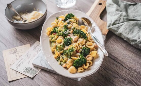 Traditionelles Orecchiette-Rezept aus Apulien mit Brokkoli und weißen Bohnen. Die italienische Pasta wird auf einer weißen Servierplatte angerichtet und mit Parmesan, Vintage-Postkarten und mediterranen Details stilvoll in Szene gesetzt