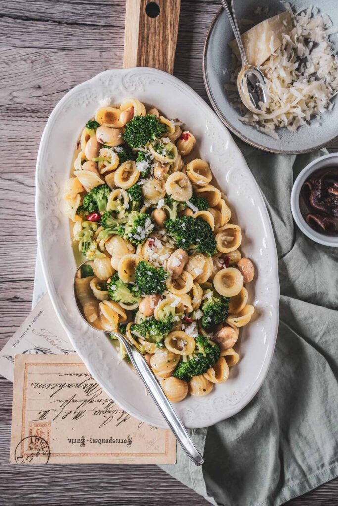 Apulisches Pastagericht mit Orecchiette, Brokkoli und weißen Bohnen auf einer weißen Porzellanplatte. Parmesan, Sardellen, Vintage-Postkarten und eine mintfarbene Serviette sorgen für mediterranes Italien-Flair auf dem Holztisch