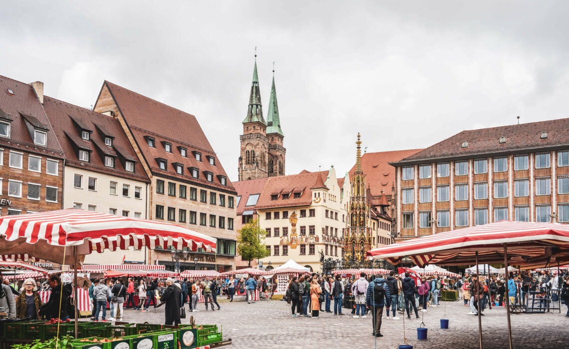 Im Herzen der Stadt: Wochenmarkt auf dem Nürnberger Hauptmarkt mit Marktständen mit rot-weiß gestreiften Planen. Im Hintergrund die Kirche St. Sebald und der Schöne Brunnen
