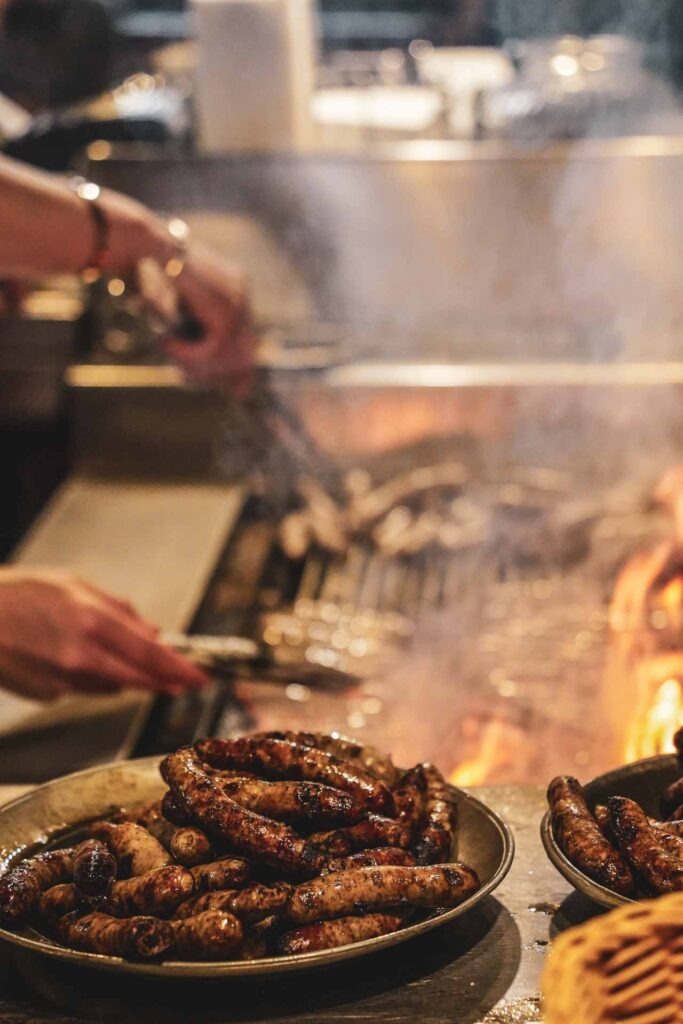 Der offene Buchenholzgrill in der Wirtsstube im Bratwursthäusle bei St. Sebald in Nürnberg, auf dem gerade frische Nürnberger Bratwürste auf offener Flamme gegrillt werden. Die fertigen Bratwürste werden auf Zinntellern im Vordergrund gestapelt