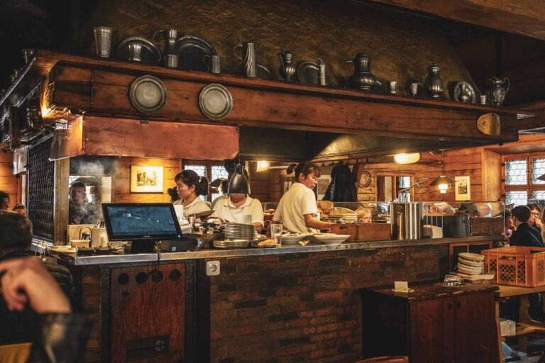 Foodguide Nürnberg – kleiner Bratwurstguide: Der große Buchenholzgrill in der holzverkleideten Wirtsstube des Bratwursthäusle bei St. Sebald in Nürnberg. Auf dem Sims sind traditionelle Zinnteller und -krüge zu sehen. Die Servicedamen am Grill tragen weiße Polo-Shirts