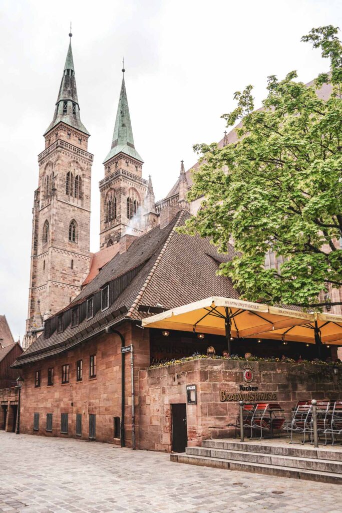 Foodguide Nürnberg – kleiner Bratwurstguide: Das historische Bratwursthäusle am Fuße der Kirche St. Sebald aus rotem Sandstein mit orangefarbenen Sonnenschirmen auf de Terrasse