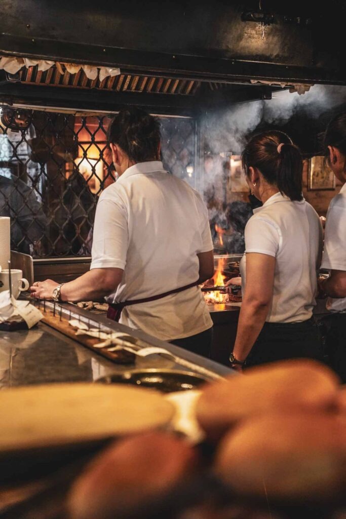 Foodguide Nürnberg - kleiner Bratwurstguide: Das Serviceteam in weißen T-Shirts im Bratwursthäusle bei St. Sebald in Nürnberg, die am offenen Grill in der Mitte der Wirtsstube stehen und Nürnberger Bratwürste grillen