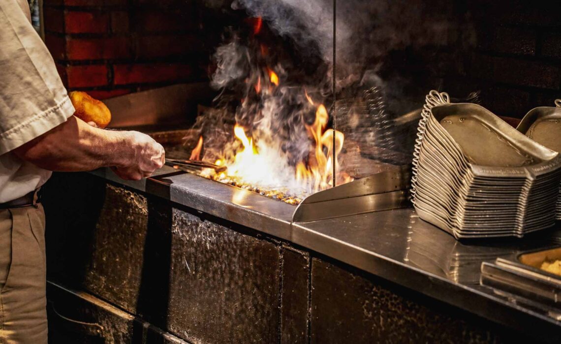Foodtipps Nürnberg: Kleiner Bratwurstguide: Offener Grill im Bratwurstglöckle im Handwerkerhof Nürnberg, auf dem gerade Bratwürste gegrillt werden. Daneben sind traditionelle Zinnteller in Glockenform zu sehen.