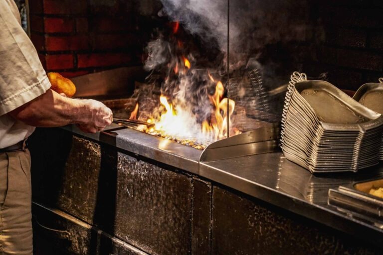 Foodtipps Nürnberg: Kleiner Bratwurstguide: Offener Grill im Bratwurstglöckle im Handwerkerhof Nürnberg, auf dem gerade Bratwürste gegrillt werden. Daneben sind traditionelle Zinnteller in Glockenform zu sehen.