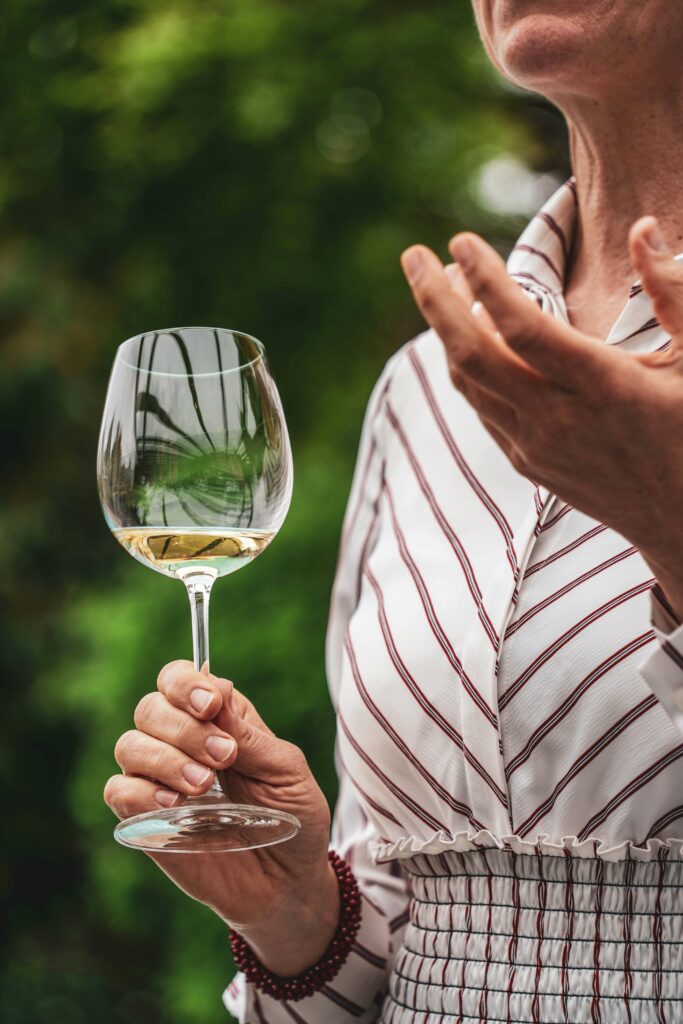Sommelière Verena Herzog von der Rotkäppchen-Mumm-Academy stellt Riesling-Weißweine von Markgraf von Baden und Chateau St. Michelle in Johann Lafers Outdoor-Küche in Guldental vor. Auf dem Foto ist die Nahaufnahme eines Glases mit Weißwein zu sehen, das sie in der Hand hält, während sie ihr Weinwissen mit den Gästen teilt.
