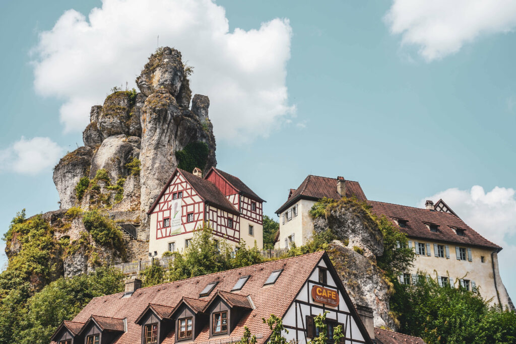 Das Bild zeigt eine Felsformation in der Fränkischen Schweiz in Oberfranken im Sommer, davor befinden sich Fachwerkhäuser.
