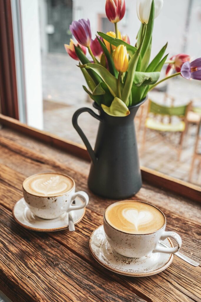 Zwei Tassen Cappuccino an einem Fensterplatz in der Kaffeerösterei BERGBRAND in der Weißgerbergasse in Nürnberg. Auf dem Tisch ist eine graue Vase mit Henkel mit bunten Tulpen zu sehen
