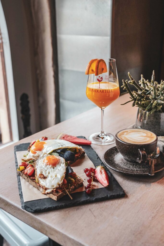 Frühstück im Café-Restaurant ZEIT & RAUM in Nürnberg: geröstete vegetarische und vegane Landbrote mit Aufstrichen, Grillgemüse, Sprossen und Spiegelei auf einem Schieferbrett. Daneben steht eine Tasse mit Capuccino und ein Glas mit frisch gepresstem Orangensaft