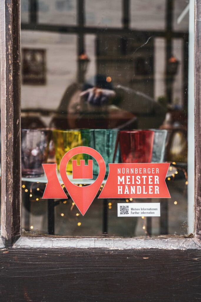 Label auf einem Ladenfenster im Nürnberger Handwerkerhof in rot mit der Aufschrift in weiß „Nürnberger Meister Händler“. Im Schaufenster sind bunte Gläser zu sehen