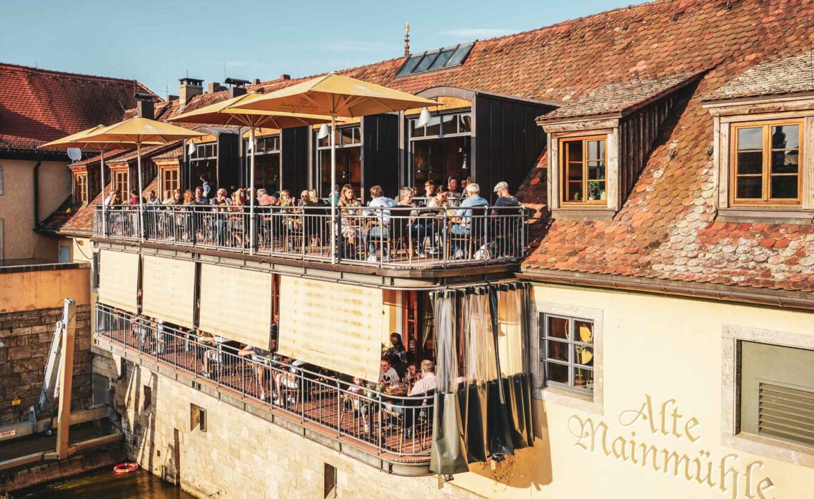 Foodtipps Würzburg: Das traditionelle fränkische Gasthaus Alte Mainmühle mit Balkonen direkt über dem Fluss Main in der Abendsonne im Sommer