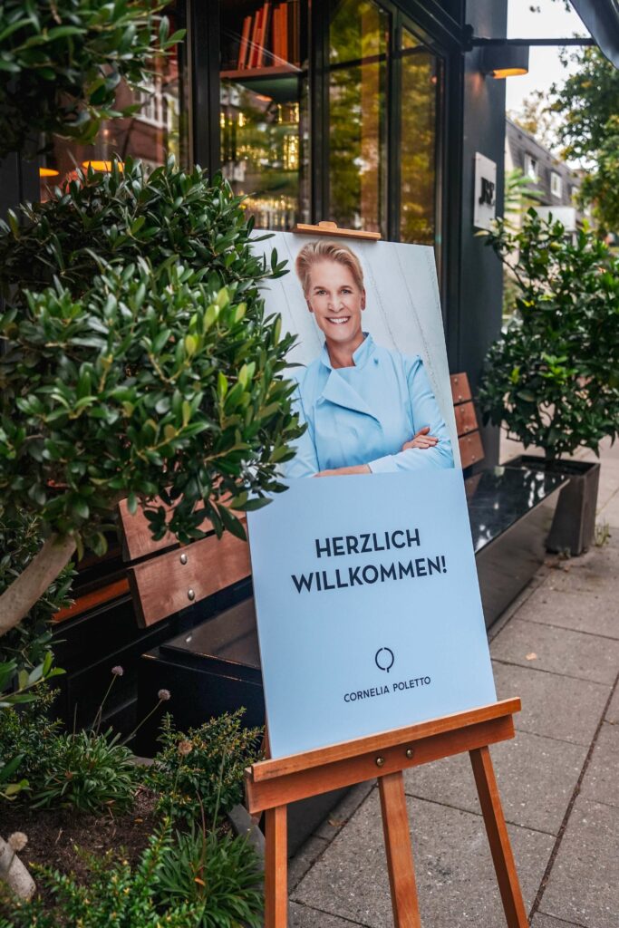 Staffelei mit Schild in Hellblau vor der Cucina Cornelia Poletto in Hamburg mit Foto der Starköchin und dem Titel: "Herzlich Willkommen" - Cornelia Poletto.