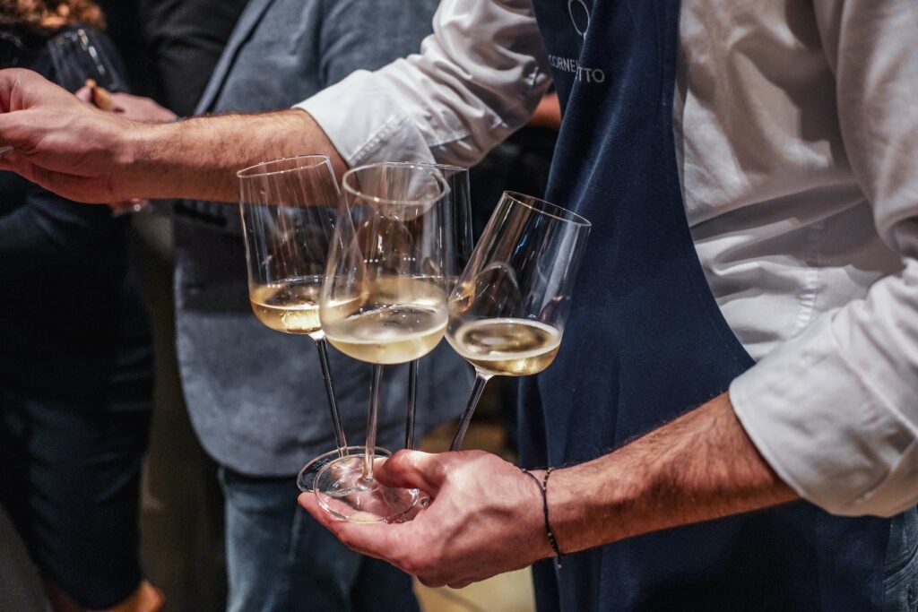 Piper-Heidsieck Champagner Blanc de Noirs »Essentiel« in Gläsern, deie vom Servicepersonal der Cucina Cornelia Poletto in marineblauen Schürzen und weißen Hemden gereicht werden
