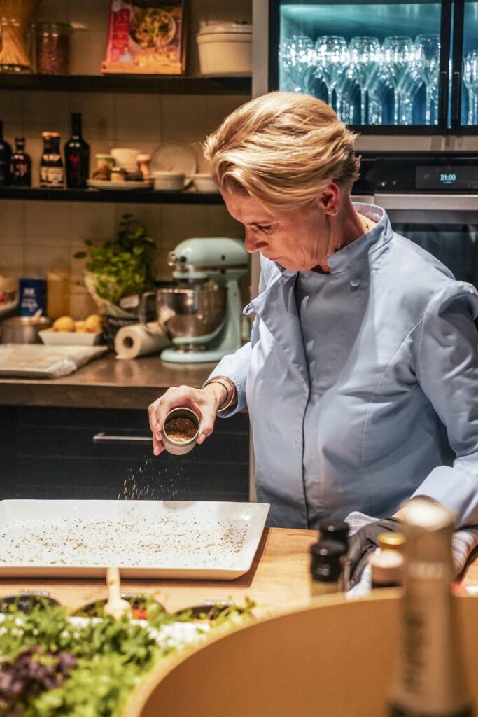 Cornelia Poletto Hamburg, die eine blaue Kochjacke trägt, streut Gewürze auf einen Teller in ihrer Kochschule in Hamburg