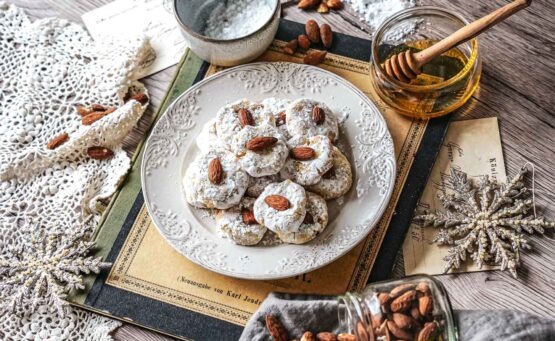 Paste di Mandorla, weiche sizilianische Mandelkekse, auf einem Teller, umgeben von Mandeln, Honig, Tannenzweigen und weihnachtlicher Dekoration auf einem Vintage-Buch