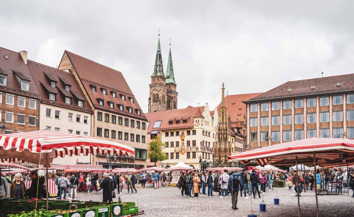 Reisetipps Nürnberg: Im Herzen der Stadt: Wochenmarkt auf dem Nürnberger Hauptmarkt mit Marktständen mit rot-weiß gestreiften Planen. Im Hintergrund die Kirche St. Sebald und der Schöne Brunnen