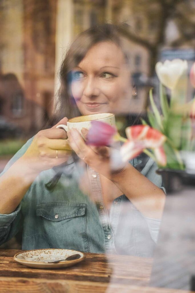 Alexandra von Tables & Fables sitzt an einem Fensterplatz im in der Kaffeerösterei BERGBRAND und blickt auf Weißgerbergasse in Nürnberg. Im Vordergrund ist eine graue Vase mit Henkel mit bunten Tulpen darin zu sehen. Alexandra trägt ein blaues Jeanshemd und hält eine Tasse mit Cappuccino in der Hand