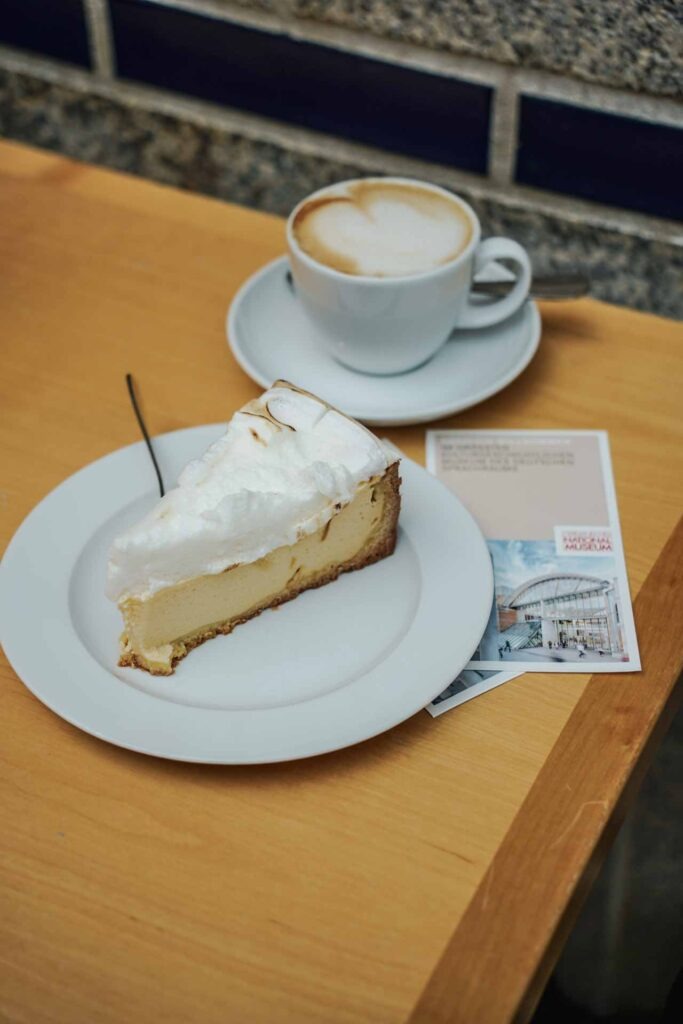 Foodtipps Nürnberg: Käsekuchen mit Baiser-Haube und Cappuccino auf einem Holztisch im Café ARTE in Nürnberg. Daneben liegt ein Flyer und Gebäudeplan des Germanischen Nationalmuseums Nürnberg