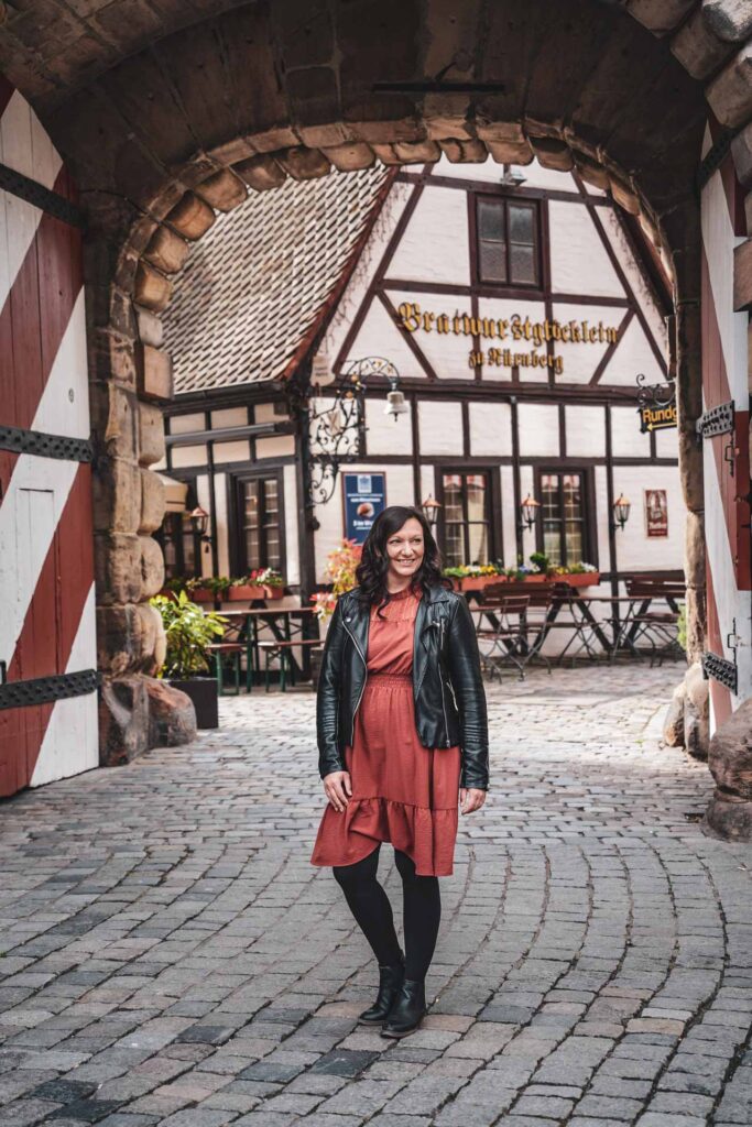 Reisetipps Nürnberg: Alexandra von Tables & Fables. Sie trägt ein kaminrotes Kleid, eine schwarze Lederjacke und Stiefel und steht am Eingang des Nürnberger Handwerkerhofs. Im Hintergrund ist das kleine Fachwerkhaus des Restaurants Bratwurstglöcklein zu sehen