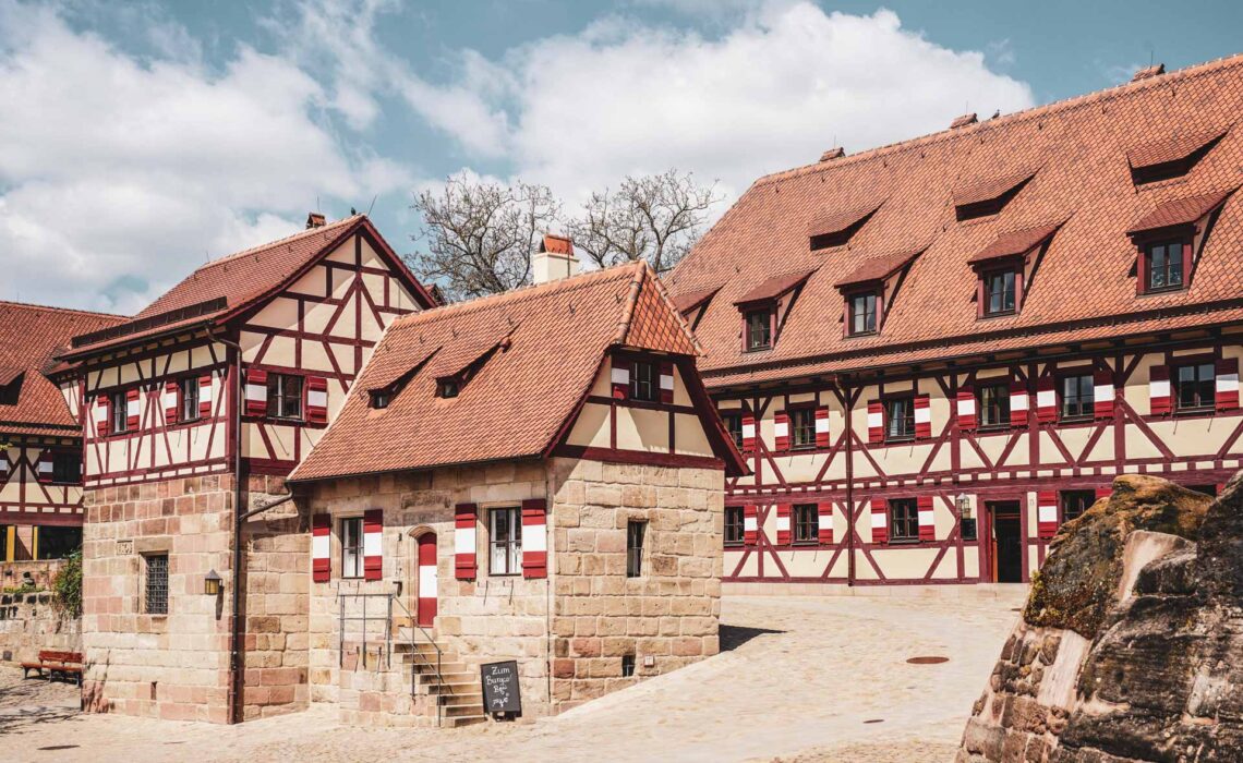 Sehenswürdigkeiten Nürnberg: Auf der Kaiserburg Nürnberg: Fachwerkhäuser mit Sandstein und typischen Fensterläden in Rot und Weiß. Im Vordergrund das Brunnenhaus der Kaiserburg