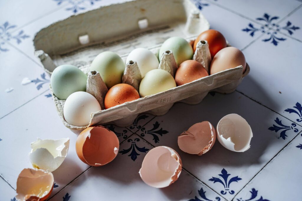 Bunte Eier zu Ostern: Diese Eier kommen schon in Pastellgrün und Pastellblau vom Bauernhof – ganz ohne Bemalen! Für den saftigen Karottenkuchen haben wir ein paar aus der Eierschachtel benutzt, die Schalen liegen daneben auf den weißen Fliesen mit dunkelblauem Muster im Sonnenlicht.