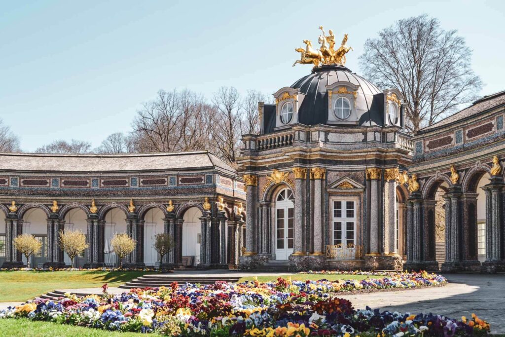 Der Sonnentempel mit Orangerie in der Eremitage Bayreuth im Frühling bei strahlendem Sonnenschein. Im Vordergrund sind Beete mit bunten Frühlingsblumen zu sehen