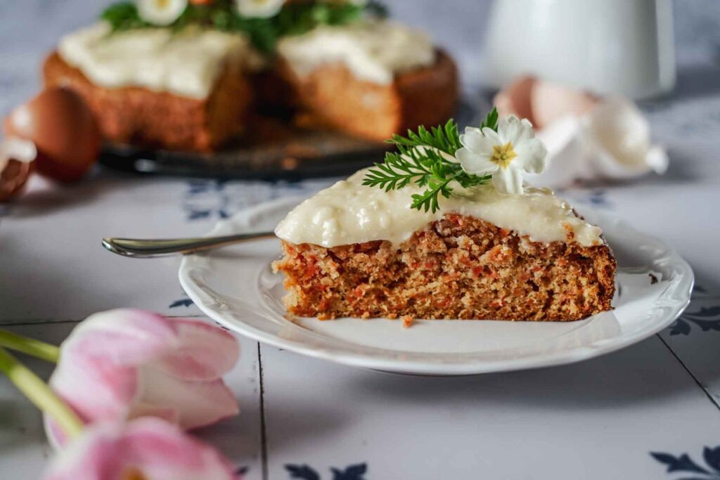 Ein Stück saftiger Karottenkuchen auf einem Teller mit Frischkäse-Buttercreme, Möhrengrün und hellgelben Frühlingsprimeln verziert. Im Vordergrund liegen hellrosafarbene Tulpen auf den weißen Fliesen mit dunkelblauem Muster. Im Hintergrund ist eine weiße Kanne mit Henkel zu sehen sowie der ganze Karottenkuchen aus einer Springfrom