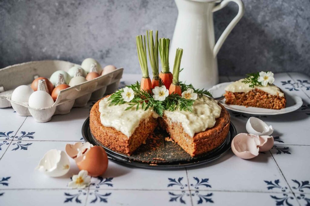 Saftiger Karottenkuchen mit Frischkäse-Buttercreme, dekoriert mit Möhrenenden, Möhrengrün und hellgelben Frühlingsprimeln. Ein Stück des Kuchens ist herausgeschnitten und im Hintergrund auf einem Teller angerichtet. Im Vordergrund liegen weiße und braune Eierschalen auf weißen Fliesen mit dunkelblauem Muster. Im Hintergrund stehen eine weiße Kanne sowie ein Eierkarton mit braunen, weißen, pastellgrünen und pastellblauen Eiern