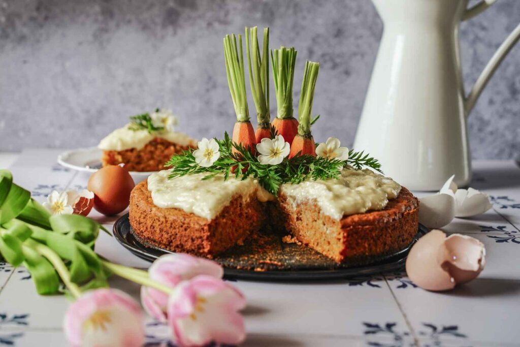 Saftiger Karottenkuchen mit Frischkäse-Buttercreme, Möhren-Enden, Möhrengrün und hellgelben Frühlingsprimeln dekoriert. Ein Stück ist herausgeschnitten und um Hintergrund auf einem Teller zu sehen. Im Vordergrund liegen hellrosafarbene Tulpen und braune Eierschalen auf den weißen Fliesen mit dunkelblauem Muster. Im Hintergrund ist eine weiße Kanne mit Henkel zu sehen.