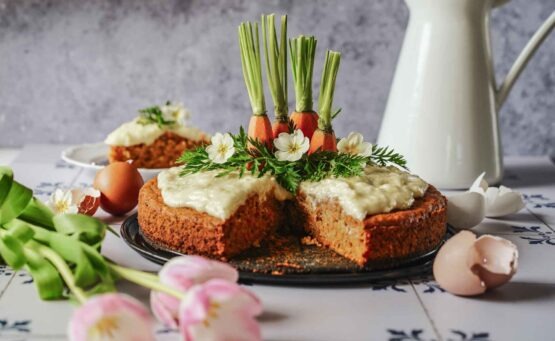 Saftiger Karottenkuchen mit Frischkäse-Buttercreme, Möhren-Enden, Möhrengrün und hellgelben Frühlingsprimeln dekoriert. Ein Stück ist herausgeschnitten und um Hintergrund auf einem Teller zu sehen. Im Vordergrund liegen hellrosafarbene Tulpen und braune Eierschalen auf den weißen Fliesen mit dunkelblauem Muster. Im Hintergrund ist eine weiße Kanne mit Henkel zu sehen.