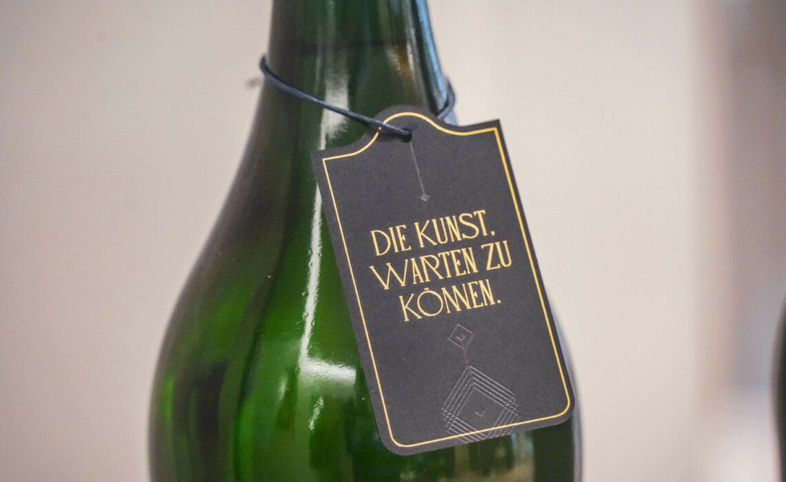 Die Kunst, warten zu können: Flaschenanhänger in Dunkelblau mit goldfarbener Aufschrift, des Slogans, der den Launch des neuen Premiumsekts begleitet