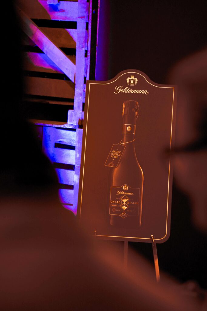 Staffelei mit Aufsteller in Dunkelblau, der die Flasche mit dem neu gelaunchten Premium-Cuvée Geldermann Grande Réserve im historischen Sektkeller bei der Präsentation zeigt. Im Hintergrund sind Holzregale zu sehen
