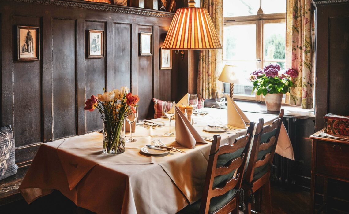 Die einladende Alte Stube im Restaurant des Romantik-Hotels Spielweg im Schwarzwald mit traditioneller Holzvertäfelung. Auf dem gedeckten Tisch stehen frische Frühlingsblumen und durch die Fenster bricht das Sonnenlicht herein
