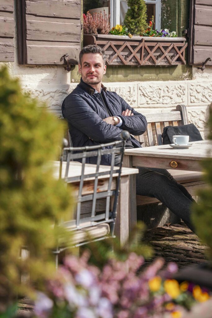 Blogger Florian von Tables and Fables auf der Sonnenterrasse des Romantik-Hotels Spielweg, umgeben von bunten Frühlingsblumen. Erträgt eine dunkelblaue Steppjacke und genießt die Frühlingssonne