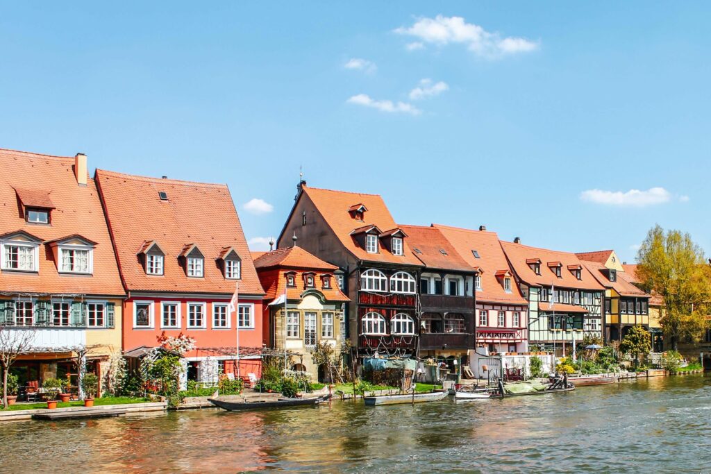 Bamberg im Frühling: Blick auf die historischen Fachwerkhäuser von Klein Venedig vom Leintritt aus. In den kleinen Gärten entlang des Regnitzufers sind farbenfrohe Frühlingssträucher und blühenden Obstbäume zu sehen.