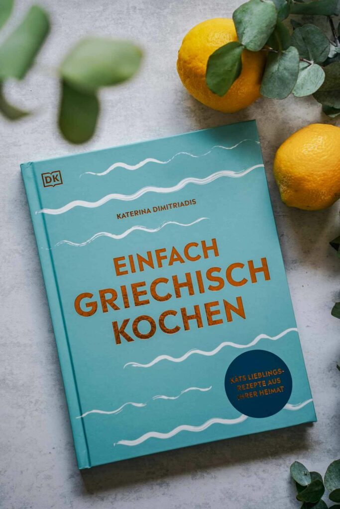 Die schönsten Kochbücher für den Sommer: Das Kochbuch Einfach Griechisch Kochen von Katerina Dimitriadis aus dem DK-Verlag. Das Bild zeigt das Buchcover in Türkis mit dem Buchtitel mit Kupferfolie und stilisierten Wellen in Weiß, umgeben Eukalyptus-Blättern und Zitronen