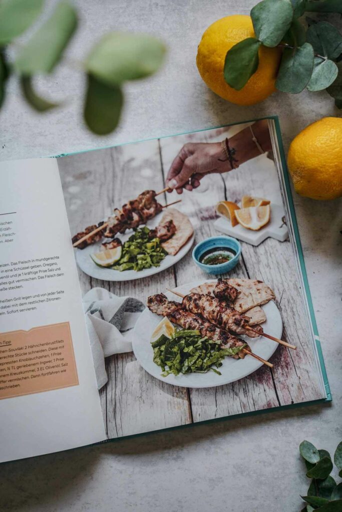 Die schönsten Kochbücher für den Sommer: Das Kochbuch Einfach Griechisch Kochen von Katerina Dimitriadis aus dem DK-Verlag. Das Bild zeigt eine Innenseite mit dem Rezeptfoto für Souvlaki mit gegrillten Fleischspießen, umgeben Eukalyptus-Blättern und Zitronen