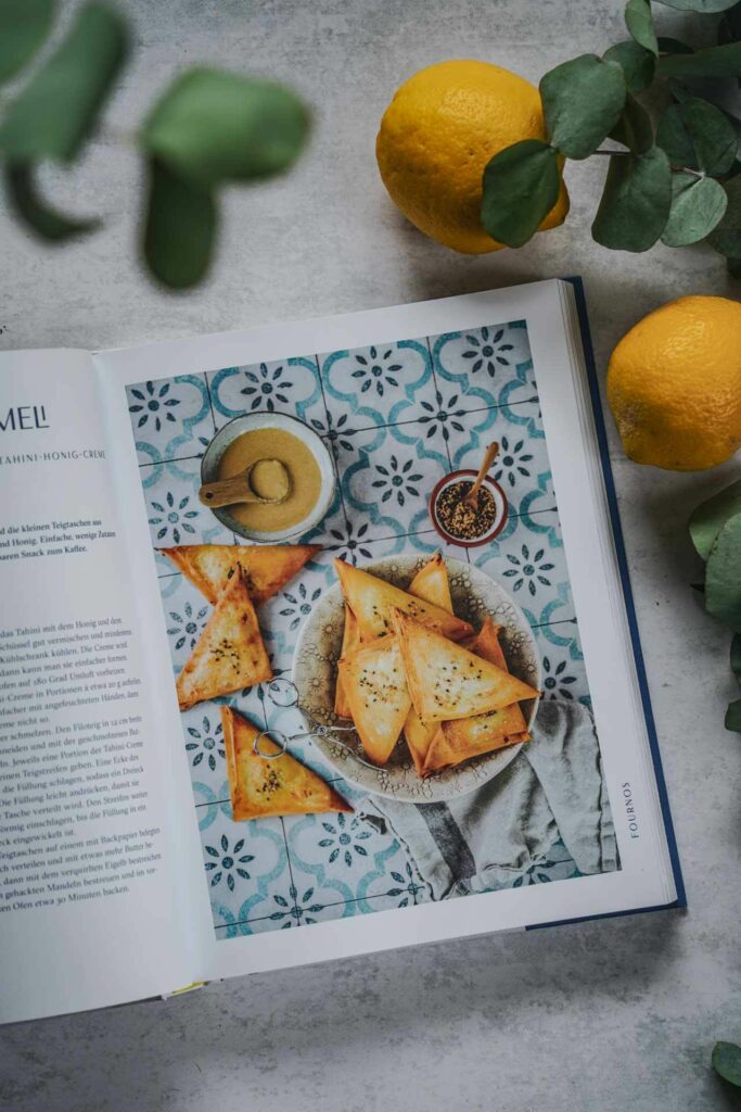 Die besten Kochbücher für den Sommer: Das Kochbuch Sonne und Meer auf dem Teller von Lazaros Kapageoroglou, erschienen im AT Verlag. Das Bild zeigt das Rezeptfoto für Tahinopitakia me Meli – Teigtaschen mit Tahini-Honig-Creme auf blauen Fliesen–, umgeben von Eukalyptus-Blättern und Zitronen