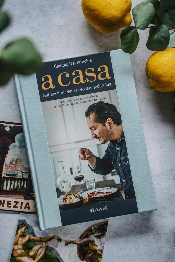 Das Italien-Kochbuch A Casa von Claudio del Principe aus dem AT-Verlag, umgeben von Vintage-Postkarten, Eukalyptus-Blättern und Zitronen. Das Bild zeigt das Buchcover in Hellblau. Darauf ist der Autor an einem gedeckten Tisch zu sehen, genussvoll ein Gericht probierend