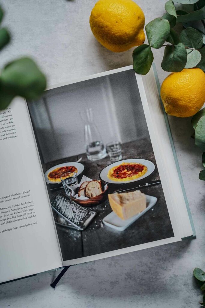 Das Italien-Kochbuch A Casa von Claudio del Principe aus dem AT-Verlag. Das Bild zeigt das aufgeschlagene Buch mit einem Rezeptfoto für das italienische Gericht Polenta e Pomodoro, umgeben von Eukalyptus-Blättern und Zitronen