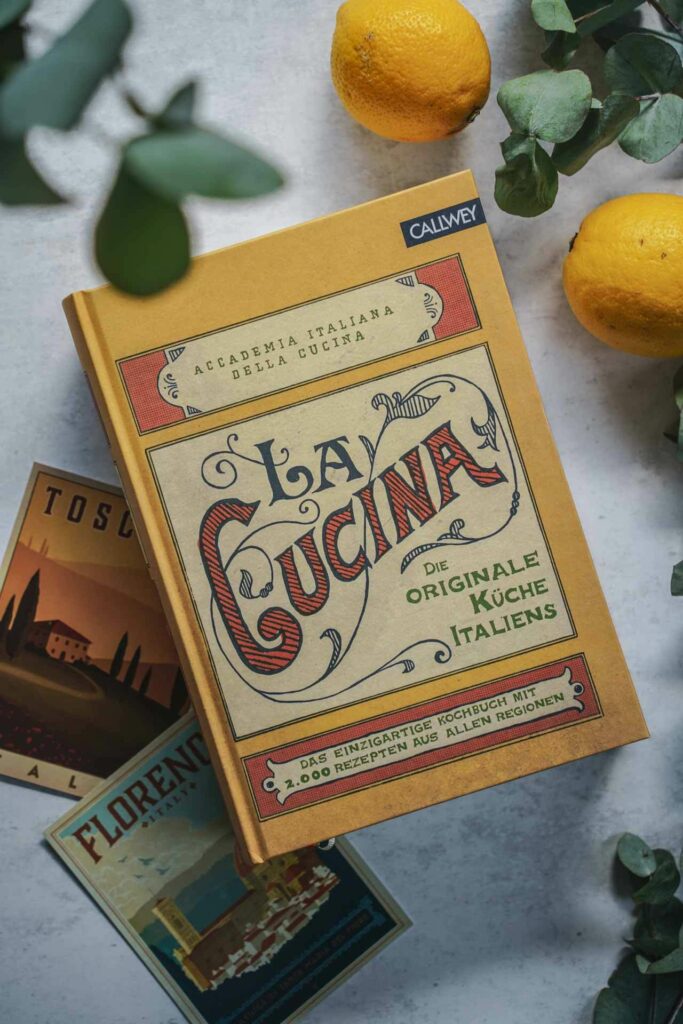 Das Italien-Kochbuch La Cucina aus dem Callwey-Verlag. Das Bild zeigt das gelbe Buchcover mit dem kunstvoll geletterten Titel in Orange, umgeben von Vintage-Postkarten, Eukalyptus-Blättern und Zitronen