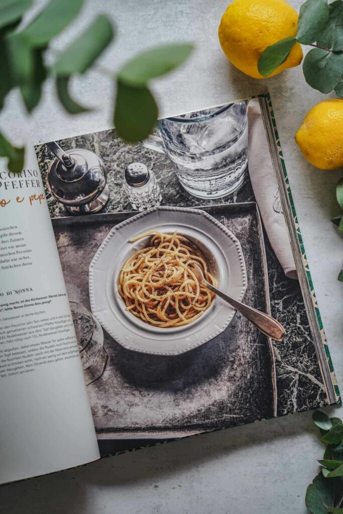 Das Italien-Kochbuch Roma in Cucina von Deborah Ferrini-Kreitmair aus dem EMF-Verlag. Das Bild zeigt das Rezeptfoto für das traditionelle römische Pastagericht Spaghetti Cacio e Pepe, umgeben von Eukalyptus-Blättern und Zitronen