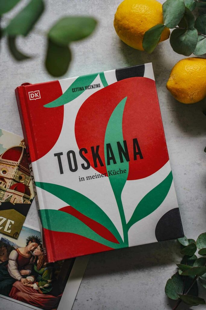 Das Italien-Kochbuch Toskana von Cettina Vicenzino aus dem DK-Verlag. Das Bild zeigt das bunt illustrierte Buchcover in Rot und Grün mit stilisierten Tomaten, umgeben von Vintage-Postkarten, Eukalyptus-Blättern und Zitronen