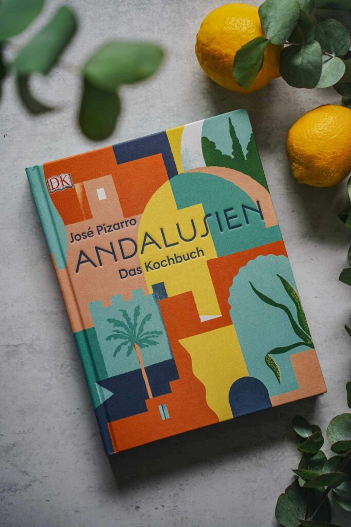 Die schönsten Kochbücher für den Sommer: Das Kochbuch Andalusien von José Pizzaro aus dem DK-Verlag. Das Bild zeigt das Buchcover in bunten Farben mit stilisierten andalusischen Häusern und Palmen, umgeben Eukalyptus-Blättern und Zitronen