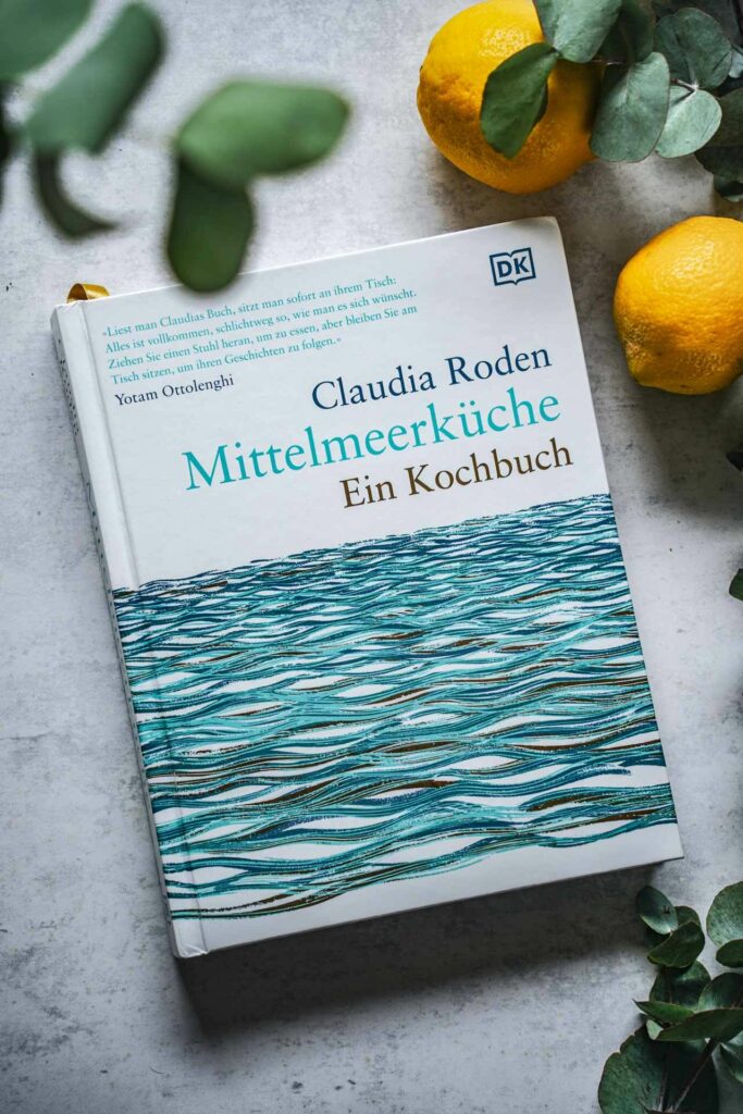 Die schönsten Kochbücher für den Sommer: Das Kochbuch Mittelmeerküche von Claudia Roden aus dem DK-Verlag. Das Bild zeigt das Buchcover mit Türkis und stilisierten Wellen, durchzogen mit Goldfolie, umgeben Eukalyptus-Blättern und Zitronen